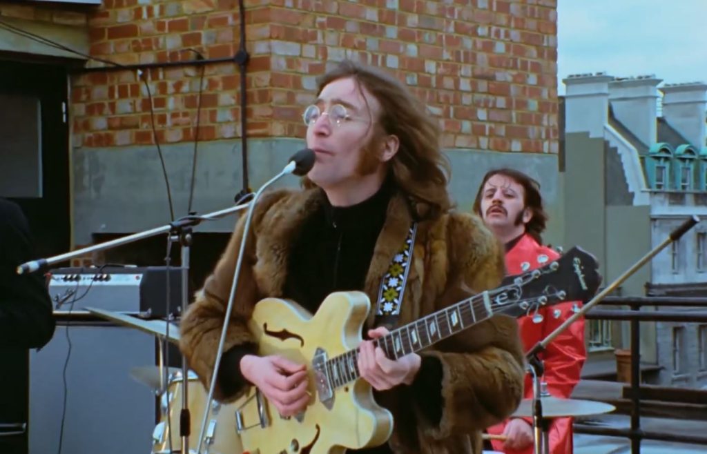 John Lennon stojący na dachu w Londynie, ubrany w futrzany płaszcz, grający na gitarze Epiphone Casino